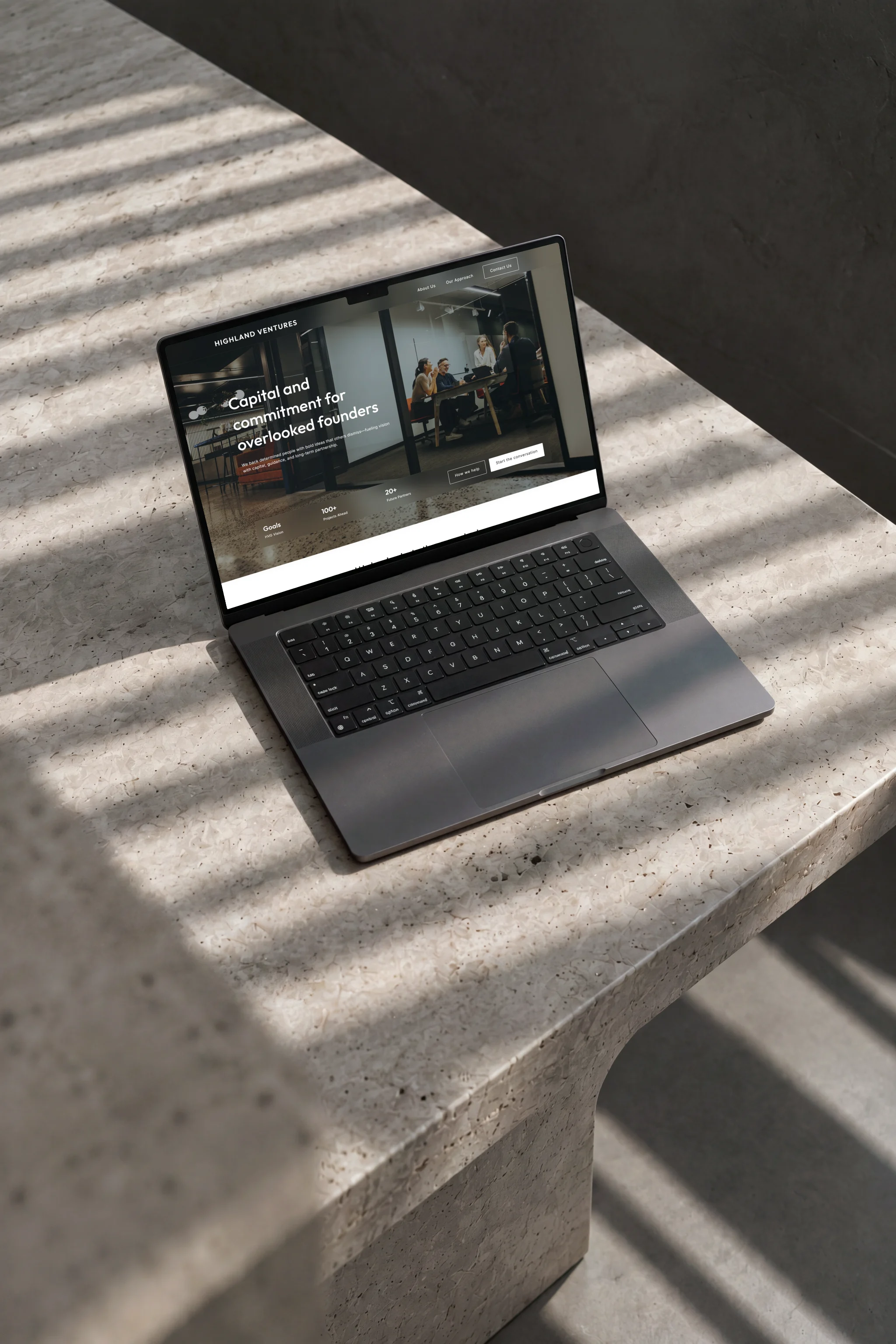 The Highland Ventures homepage on a black laptop on the corner of a pale stone table, striped window light across it